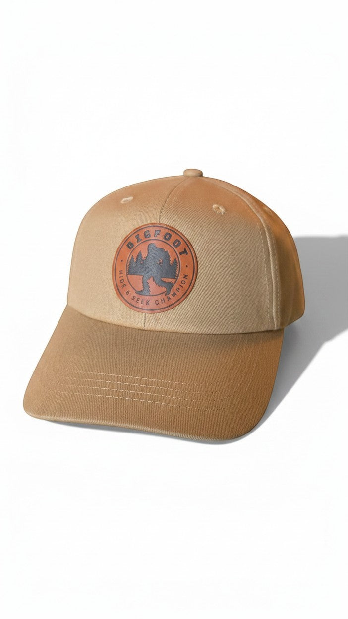 Moove Headwear tan trucker hat with a round leather patch featuring Bigfoot silhouette and trees. Adjustable fit.