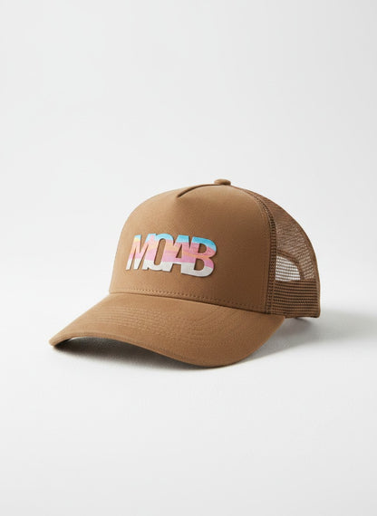 Moove Headwear brown suede trucker hat with colorful MOAB graphic, mesh back, and adjustable strap. Desert landscape background.