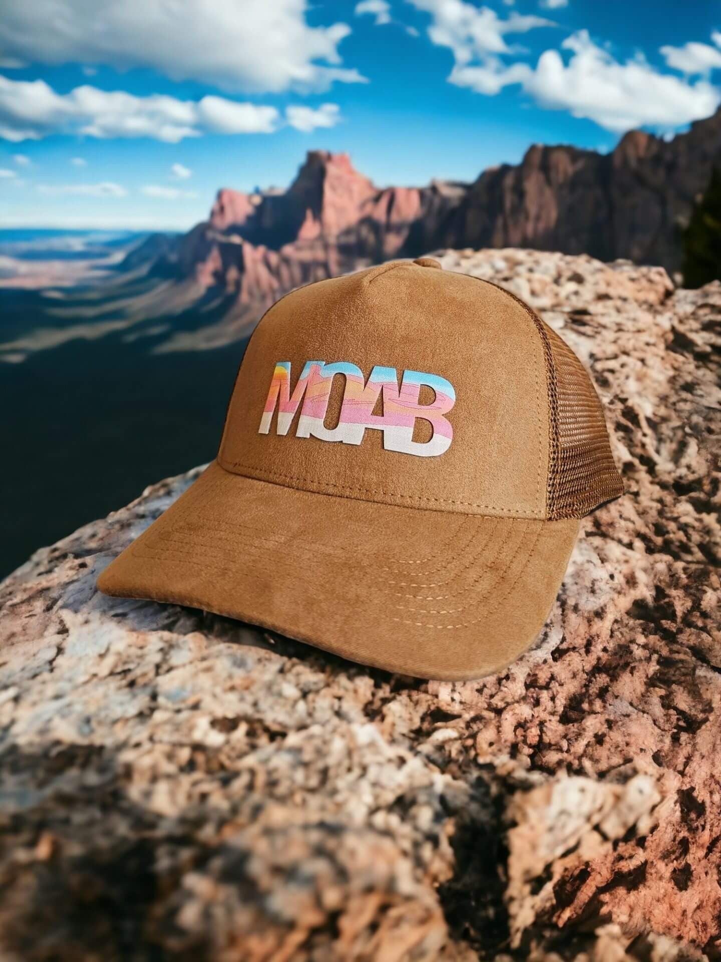 Moove Headwear brown suede trucker hat with colorful MOAB graphic, mesh back, and adjustable strap. Desert landscape background.
