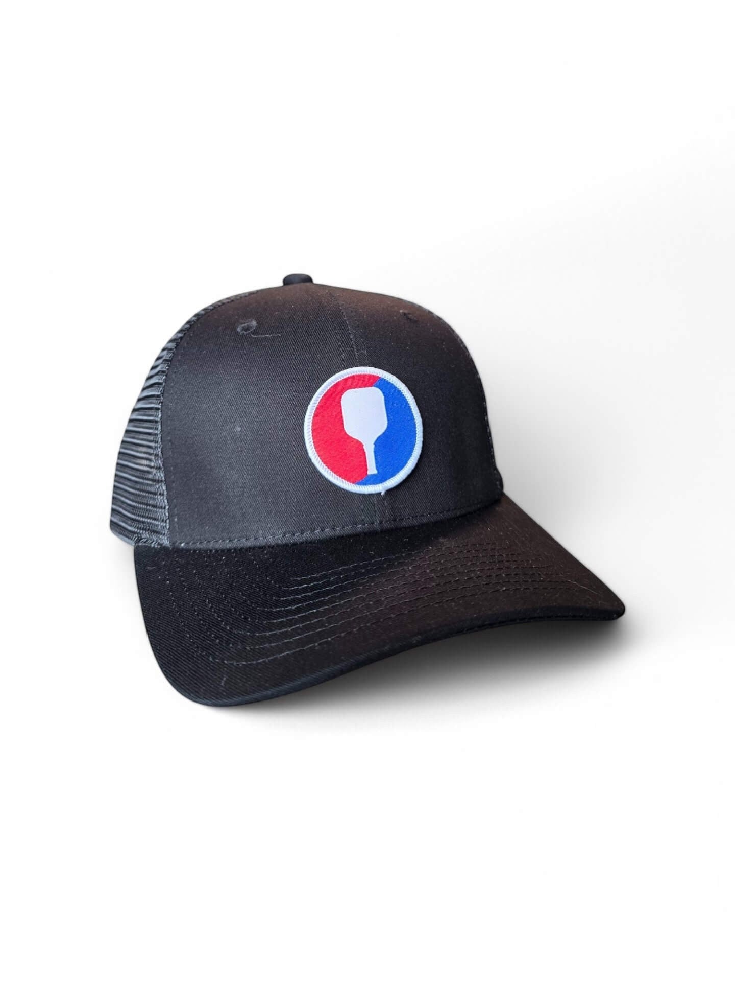 Moove Headwear black trucker hat with mesh back and a circular pickleball paddle patch in red, white, and blue.