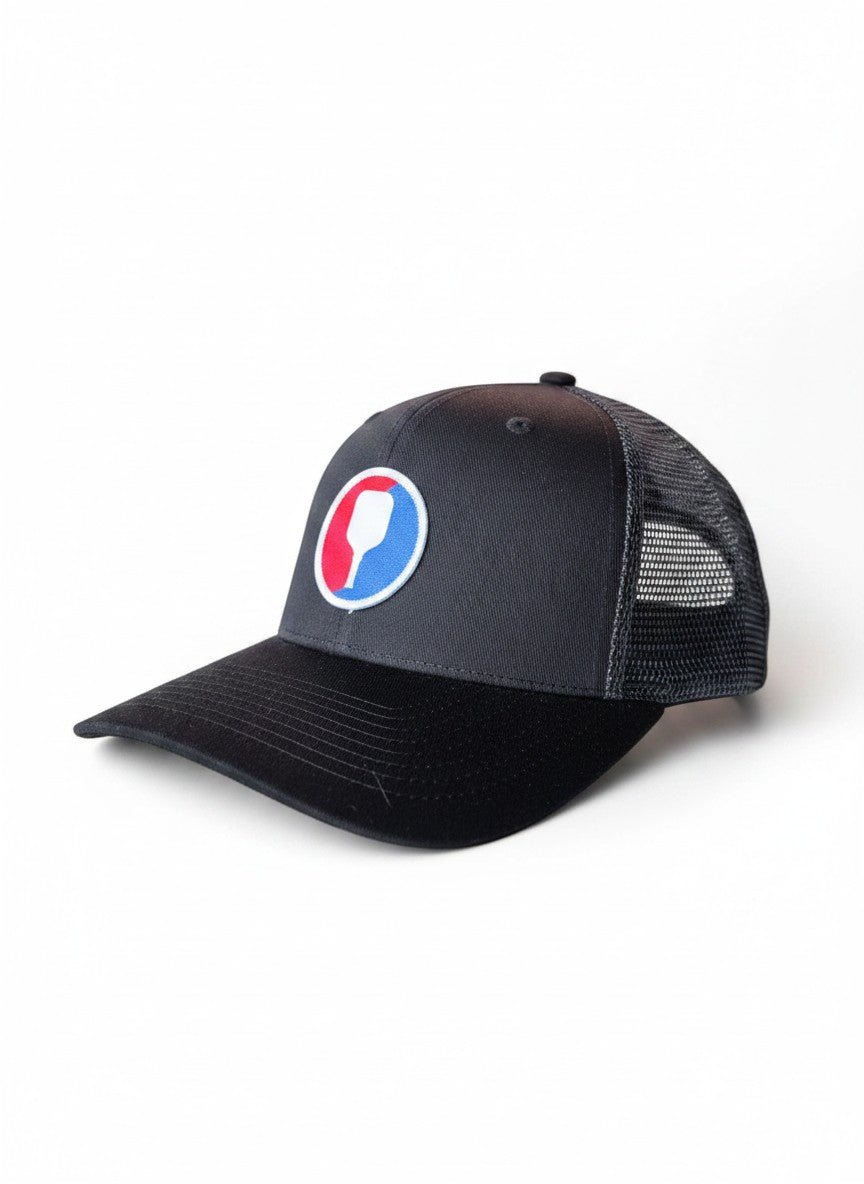 Moove Headwear black trucker hat with mesh back and a circular pickleball paddle patch in red, white, and blue.